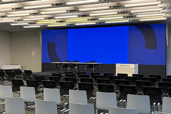 Ein großer Tagungsraum, in dem viele graue und schwarze Stühlen vor einer großen LED-Wall stehen