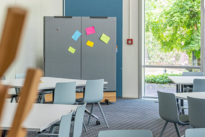 Moderner Seminarraum für Kick-off-Meetings mit flexibel angeordneten Tischen, Pinnwand mit bunten Moderationskarten und Blick ins Grüne.